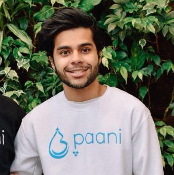 Sikander "Sonny" smiles in a white shirt with the name of his project, Paani, on it. He stands in front of a tree or bush outside