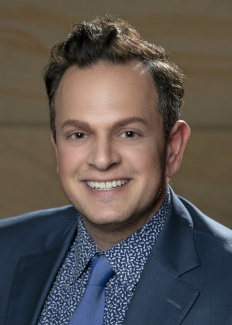 Photo of a man with brown hair smiling and wearing a gray suit with a blue shirt. 
