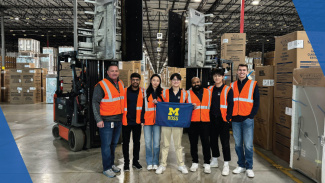 A small group of Michigan Ross students in orange and yellow safety vests pose in a warehouse filled with boxes as they hold up the blue and yellow Ross flag