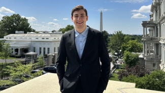 Photo of Michigan Ross BBA student Karim Kadouh standing outside of the West Wing at the White House. He is wearing a navy blue suit with a light blue shirt. He has dark hair and is looking at the camera and smiling.There are buildings in the background and the sky is really blue with a couple of clouds. 