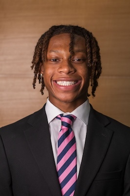 Photo of man with curly hair smiling and wearing a black suit with a white shirt with a pink and blue striped tie